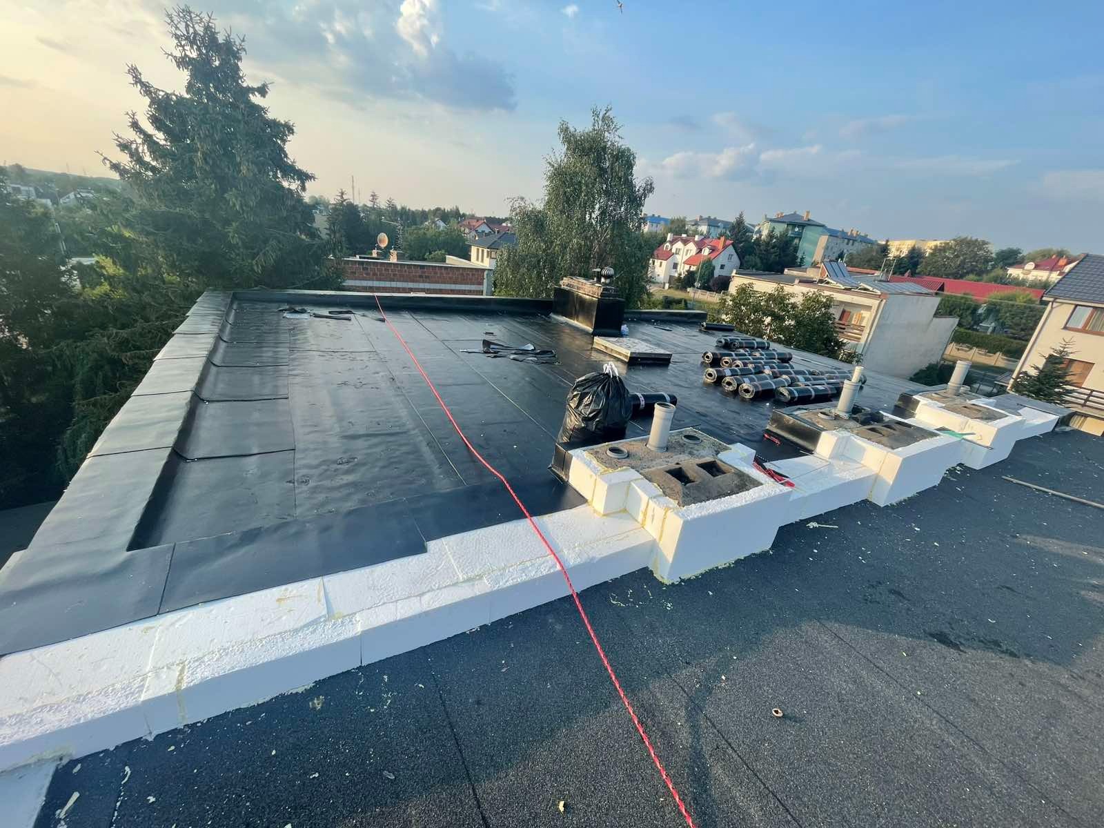 Płaski dach pokryty papą termozgrzewalną z widocznym ociepleniem styropianem. W tle zabudowa jednorodzinna i drzewa. Częściowo wykonane obróbki.