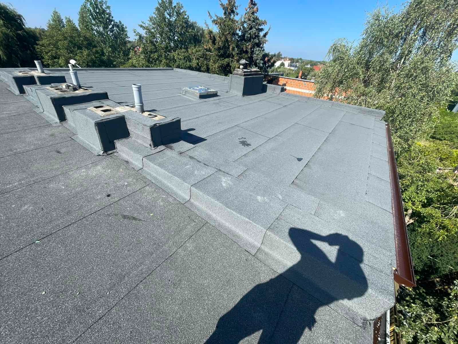 Szary dach pokryty papą termozgrzewalną z kominami wentylacyjnymi i brązową rynną. Cień osoby w prawym dolnym rogu. Widok z góry na budynek i zieleń w tle.