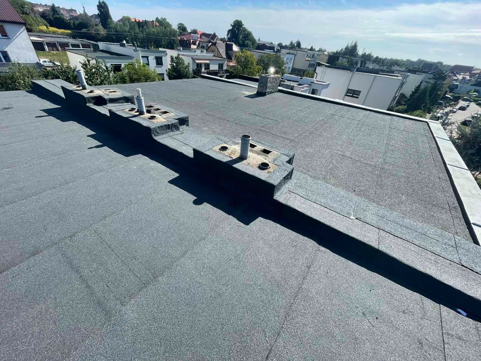 Szary, płaski dach pokryty papą termozgrzewalną z kominkami wentylacyjnymi i obróbkami blacharskimi, widok z góry na tle zabudowy miejskiej w słoneczny dzień.