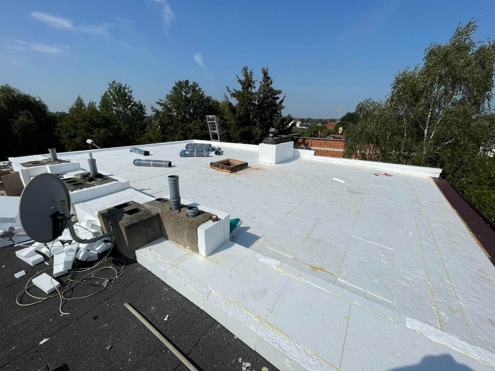 Białe płyty ociepleniowe na dachu z kominami i anteną satelitarną, widok z góry w słoneczny dzień. Częściowo widoczna drabina i zieleń w tle.