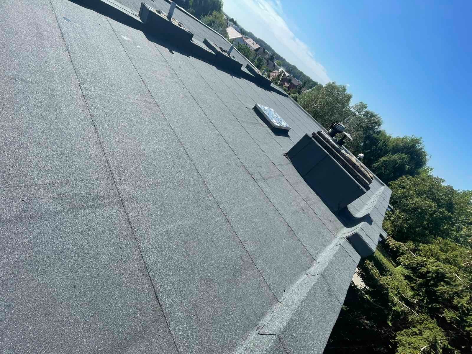 Czarny, płaski dach pokryty papą termozgrzewalną z kominem i świetlikiem dachowym. Widok z góry na tle zieleni i błękitnego nieba.