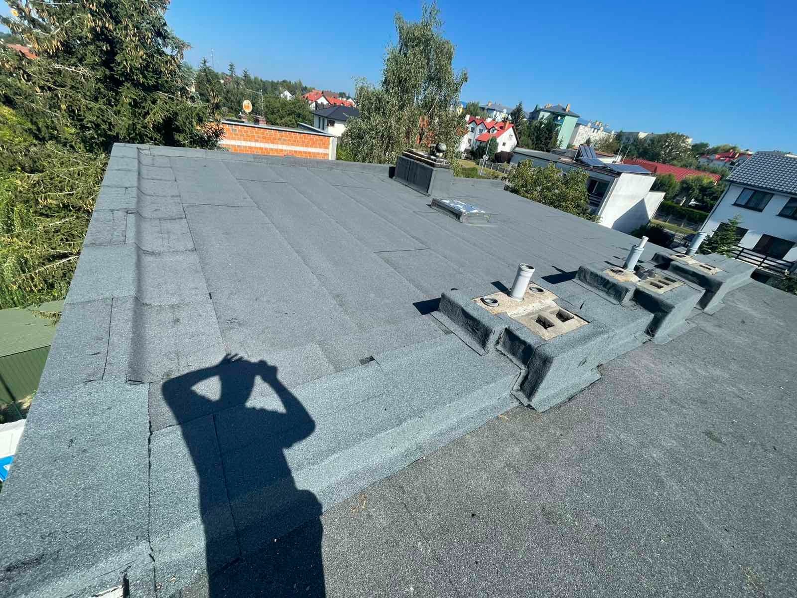 Szary, płaski dach pokryty papą termozgrzewalną z kominkami wentylacyjnymi i cieniem osoby robiącej zdjęcie na tle zabudowy miejskiej w słoneczny dzień.