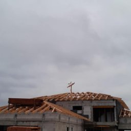 Daniel Siwak - Budowa domu z betonu komórkowego w trakcie realizacji. Widoczna konstrukcja dachu z drewnianych belek, rusztowania i materiały budowlane. Pochmurne niebo w tle.