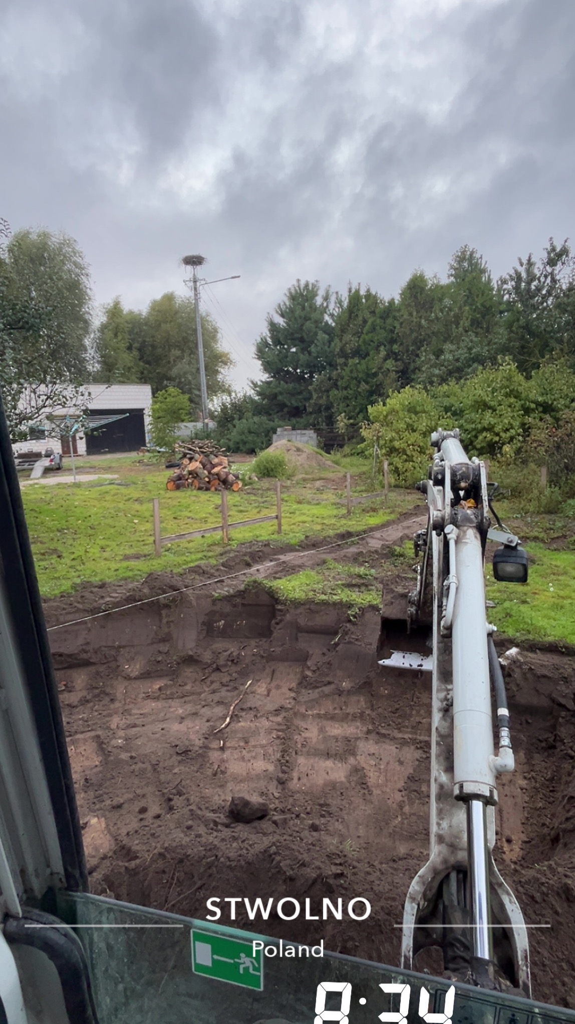 Wykopany rów w ziemi widziany z kabiny koparki, w tle drzewa i gniazdo bocianie na słupie. Prace ziemne w Stwolnie, Polska.