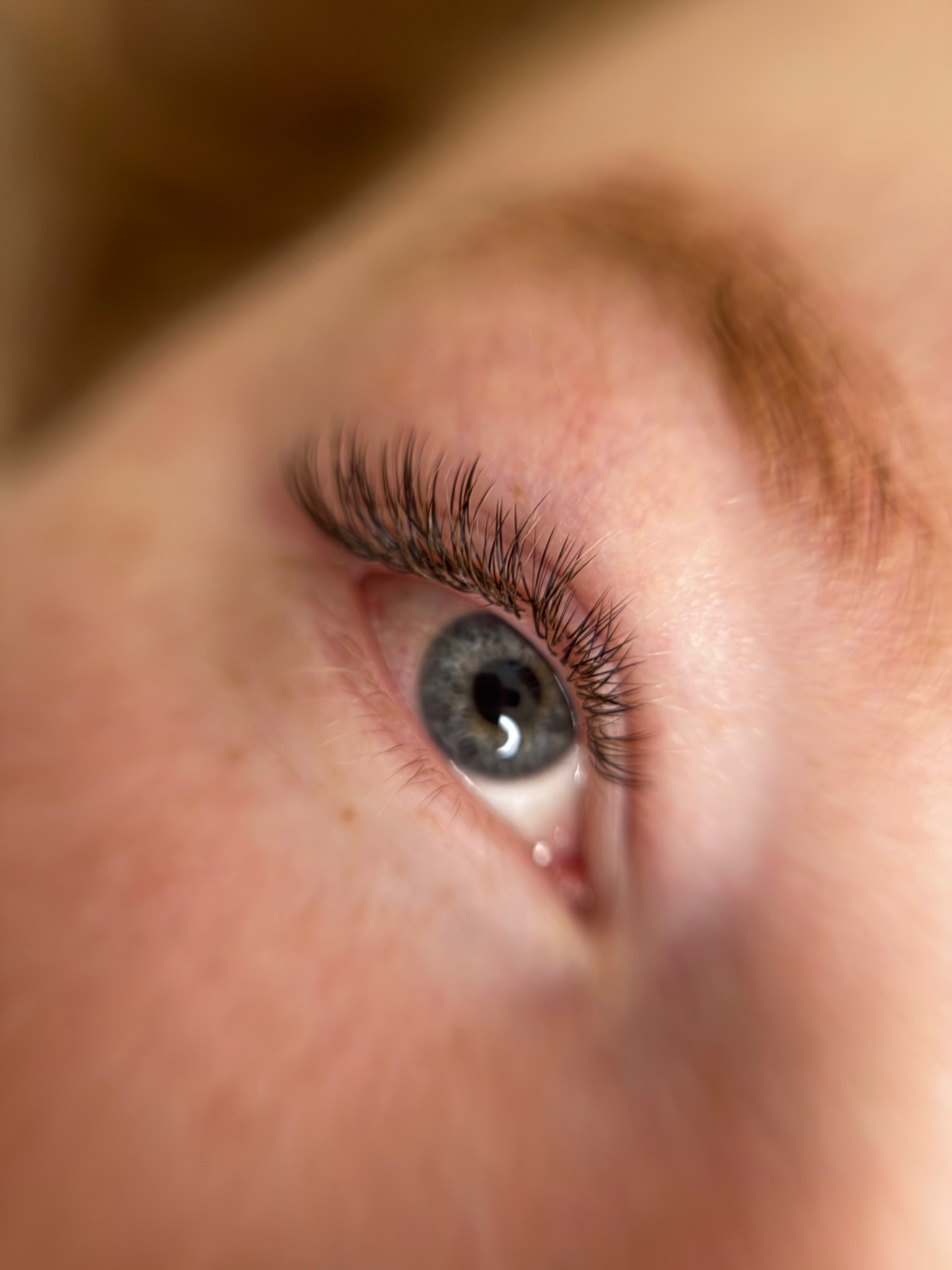 Zbliżenie na oko z efektem przedłużonych rzęs 2:1, tzw. mokry efekt. Widoczne pojedyncze, ciemne rzęsy, delikatny makijaż oka i fragment brwi.