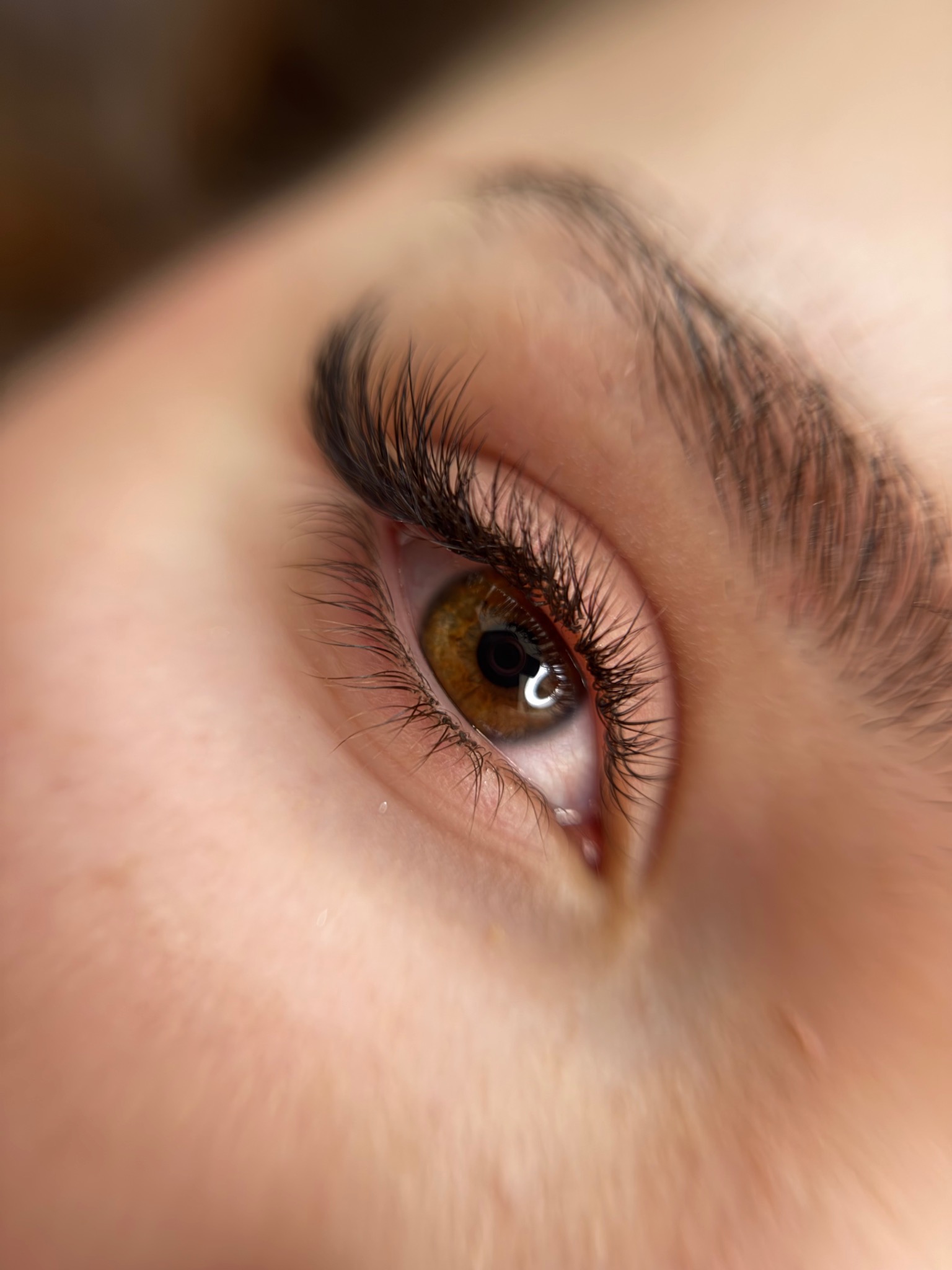 Zbliżenie na oko z brązową tęczówką i wyraźnie przedłużonymi, ciemnymi rzęsami. Widoczny delikatny blask w oku i fragment brwi. Makro, estetyczne.