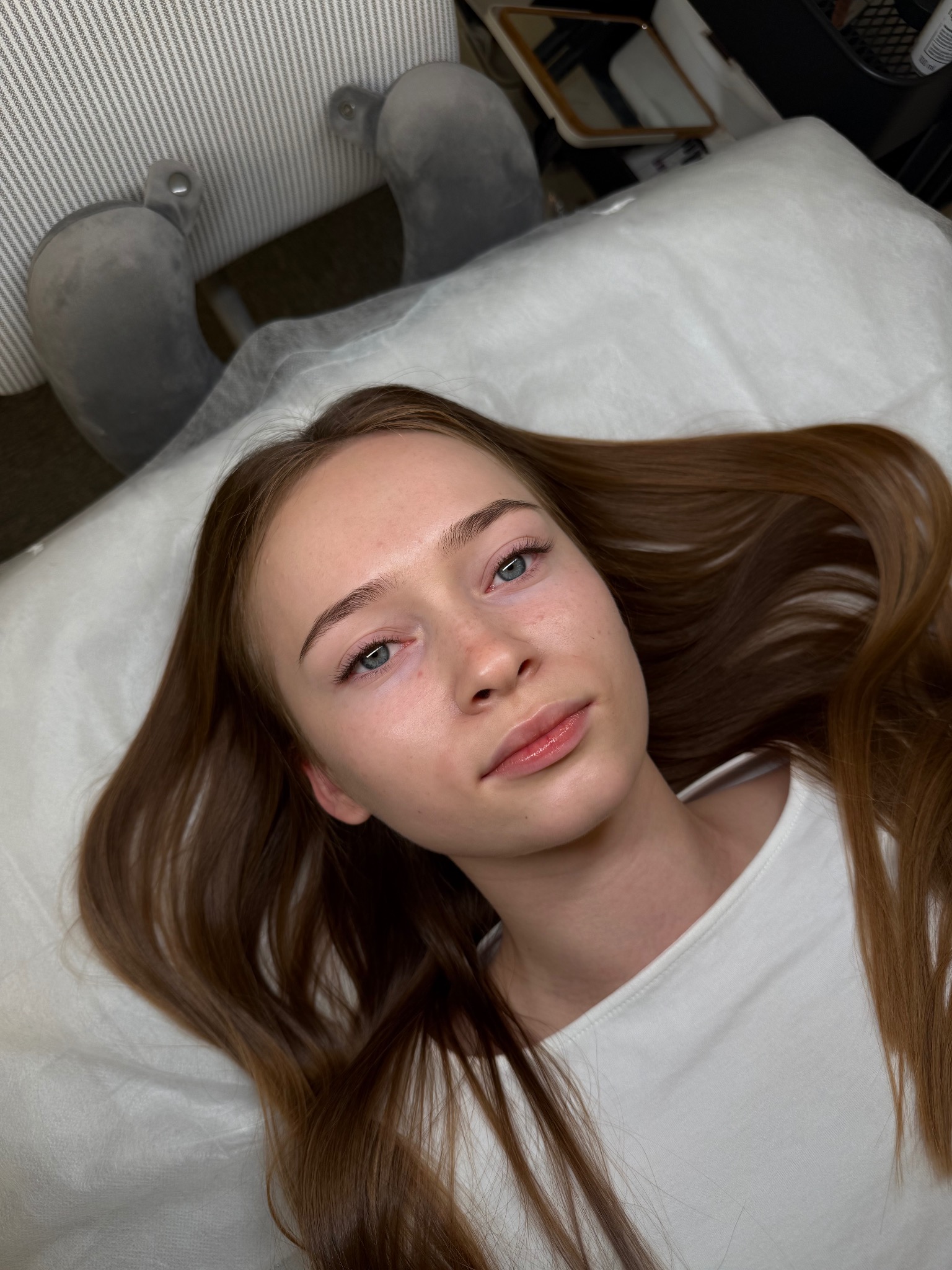 Młoda kobieta leży na leżance kosmetycznej po zabiegu przedłużania rzęs. Widoczne długie, brązowe włosy i delikatny makijaż. Jasne, naturalne światło.