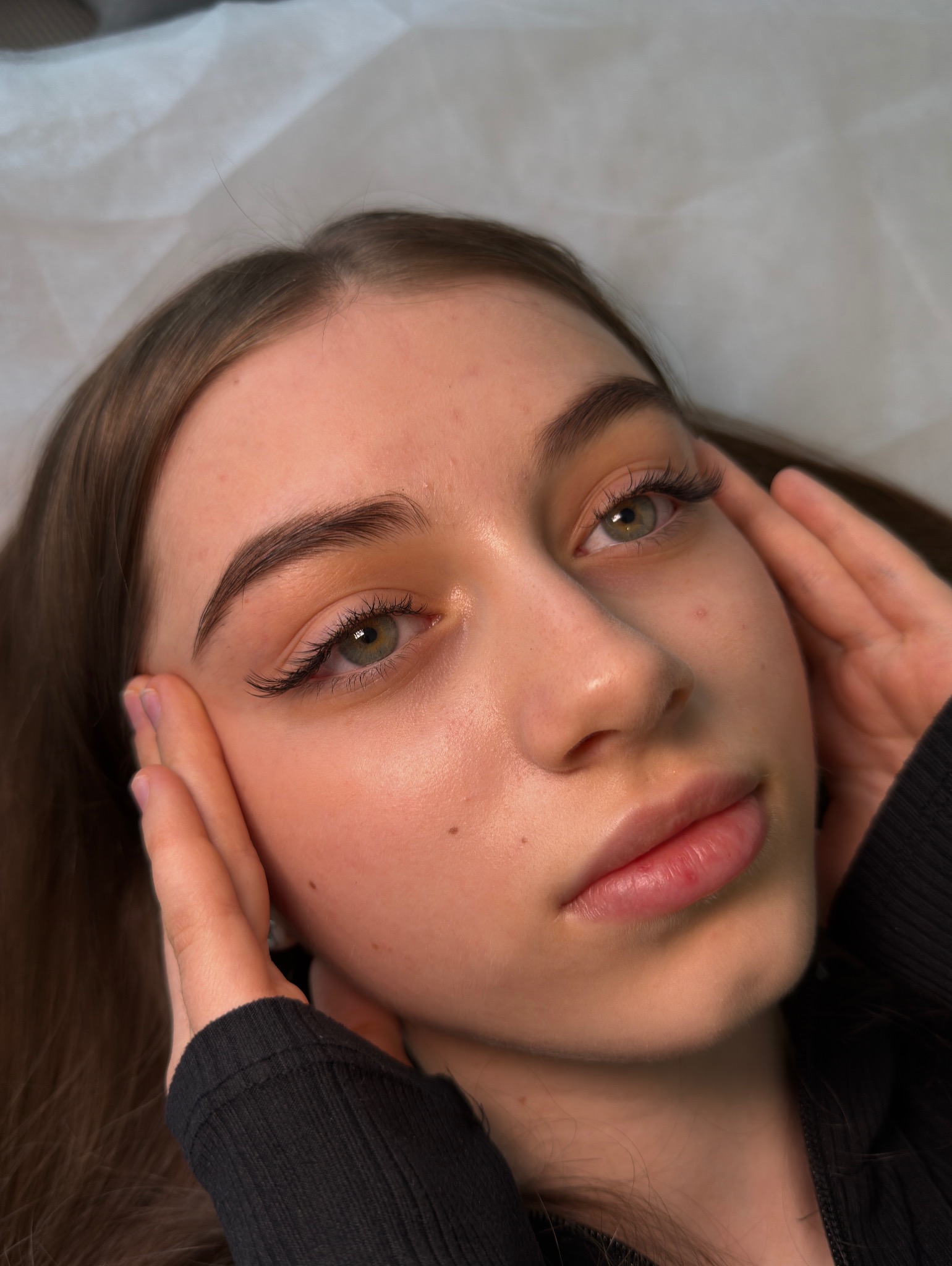 Zbliżenie na twarz młodej kobiety z profesjonalnie przedłużonymi rzęsami, dłońmi dotyka policzków. Widoczne zielone oczy i naturalny makijaż.