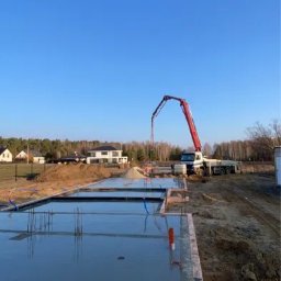 DOM-BUD - Wylewanie betonu na fundamenty domu jednorodzinnego za pomocą pompy do betonu. Widoczne szalunki, zbrojenie i świeżo wylany beton. W tle dom w budowie i błękitne niebo.