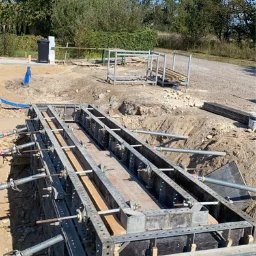 DOM-BUD - Szczegółowe ujęcie szalunku fundamentu w wykopie, z widocznymi elementami konstrukcyjnymi i zbrojeniem, na tle zieleni i błękitnego nieba.