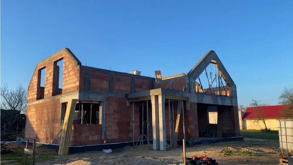 Niedokończony dom z czerwonej cegły i betonu w słoneczny dzień. Widoczne szalunki i podpory. Budowa w toku, zarys dachu i okien. Tło: inne budynki i błękitne niebo.