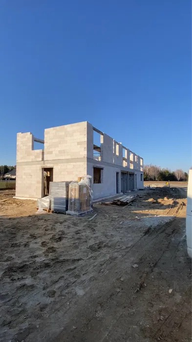 Wznoszenie ścian parterowego domu z betonu komórkowego. Częściowo wymurowane ściany z widocznymi otworami okiennymi i drzwiami. Na pierwszym planie palety z bloczkami.