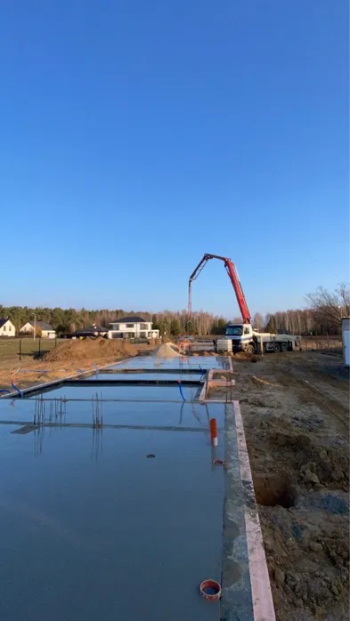 Wylewanie betonu na fundamenty domu jednorodzinnego za pomocą pompy do betonu. Widoczne szalunki, zbrojenie i świeżo wylany beton. W tle dom w budowie i błękitne niebo.