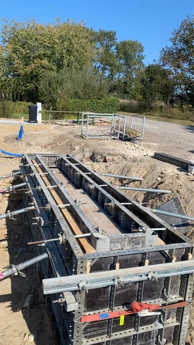 Szczegółowe ujęcie szalunku fundamentu w wykopie, z widocznymi elementami konstrukcyjnymi i zbrojeniem, na tle zieleni i błękitnego nieba.