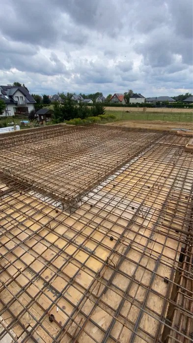 Zbrojenie stropu na deskowaniu, widok z góry. Siatka z prętów stalowych tworzy regularny wzór. W tle zabudowa jednorodzinna i pochmurne niebo.