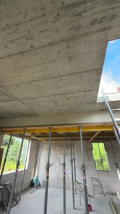 Wnętrze budynku w trakcie budowy z widocznym stropem żelbetowym podpartym metalowymi stemplami. Widoczne okna i ściany z bloczków.