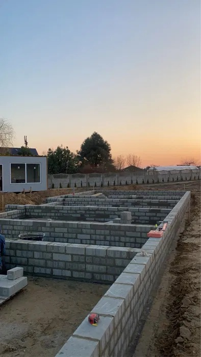 Wznoszenie fundamentów z bloczków betonowych na tle zachodzącego słońca. Widoczny zarys przyszłego budynku i mały budynek gospodarczy w tle.