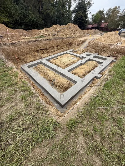 Zalany betonowy fundament pod budynek, widoczne wystające pręty zbrojeniowe. Wykop otoczony ziemią i trawą. Ujęcie z góry, w tle drzewa i częściowo widoczny samochód.
