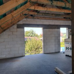 Maciej Warczak - Wnętrze domu w trakcie budowy: ściany z betonu komórkowego, drewniana konstrukcja dachu, widok na zieleń przez otwory okienne. Częściowo widoczne bloczki H+H.