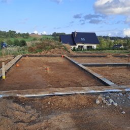 Maciej Warczak - Zalane fundamenty domu jednorodzinnego z widocznym zbrojeniem i izolacją, na tle zielonego krajobrazu i błękitnego nieba. Widoczne elementy instalacji.