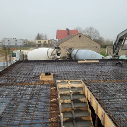 Maciej Warczak - Zbrojenie stropu z betonem w szalunku, widoczne dwa betoniarki i dźwig podający beton. W tle zabudowa wiejska i pochmurne niebo. Etap budowy domu.