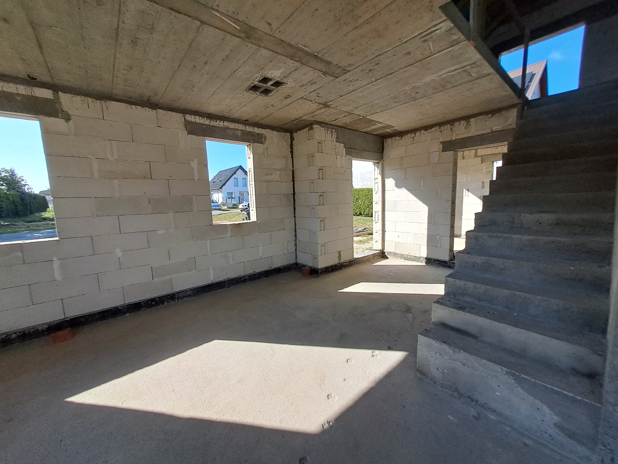 Surowy stan wnętrza domu w budowie: ściany z betonu komórkowego, betonowy strop z otworami, betonowe schody, wpadające światło słoneczne przez okna i drzwi.