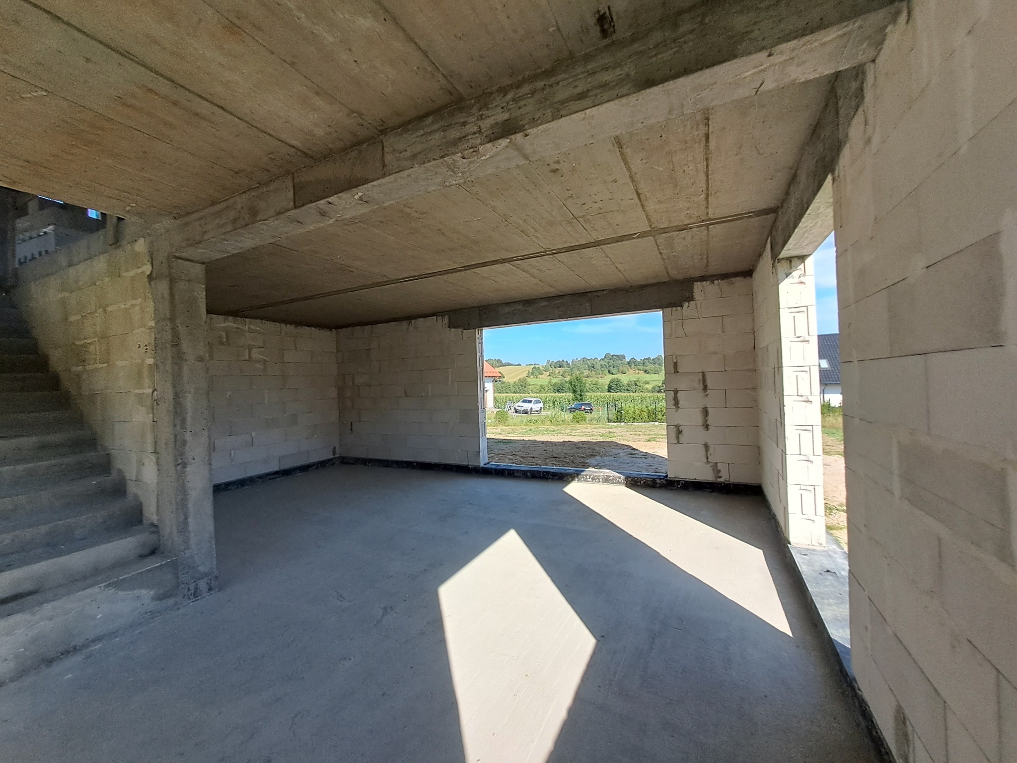 Surowy stan wnętrza domu w budowie: ściany z betonu komórkowego, betonowy strop, schody. Widok na zewnątrz przez otwór okienny, krajobraz wiejski w tle.