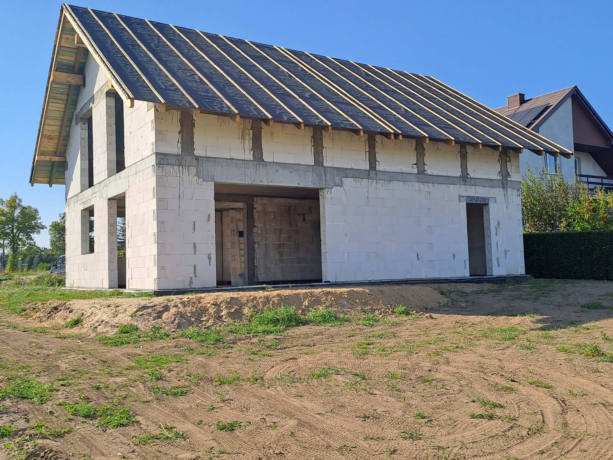 Surowy stan domu z białych bloczków, z widoczną konstrukcją dachu pokrytą czarną folią i drewnianymi łatami. Widoczne otwory okienne i drzwiowe, teren wokół w stanie naturalnym.