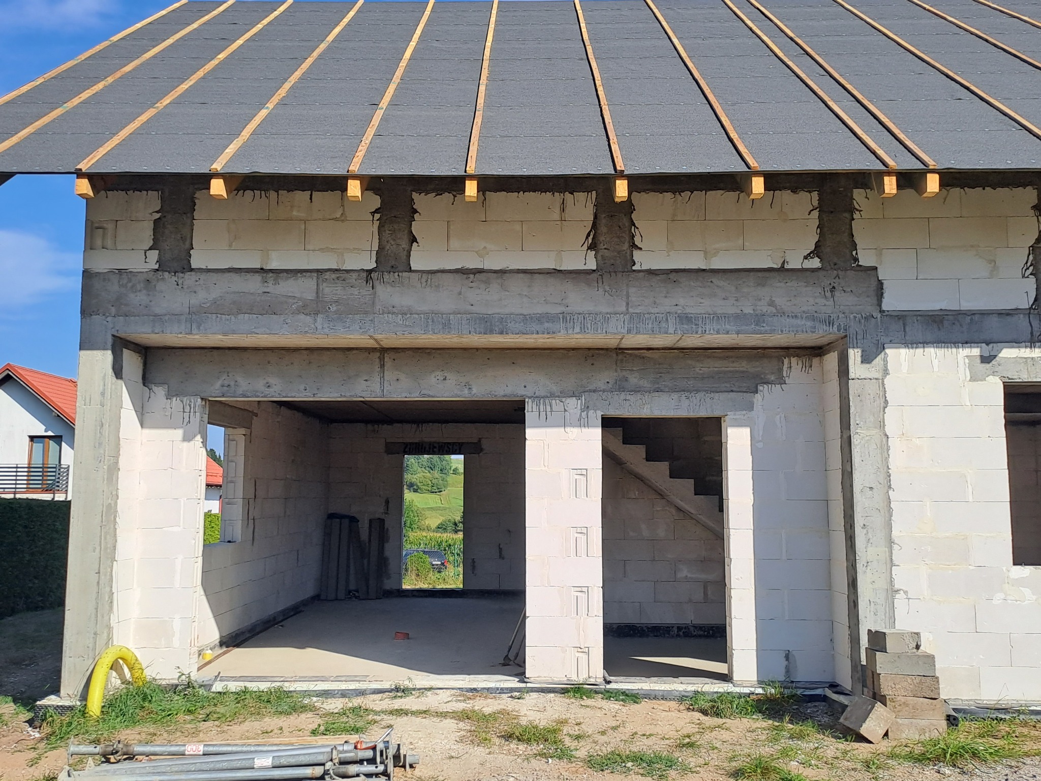 Surowy stan domu z betonu komórkowego z częściowo wykonanym dachem. Widoczne elementy konstrukcyjne i otwarte przestrzenie okienne. Na pierwszym planie narzędzia budowlane.