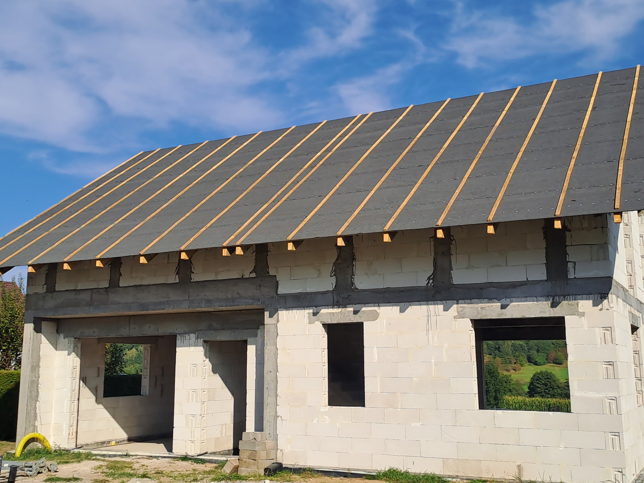 Surowy stan domu z białego bloczka, widoczne otwory okienne i drzwiowe, na dachu papa i łaty, błękitne niebo w tle. Dom w trakcie budowy.