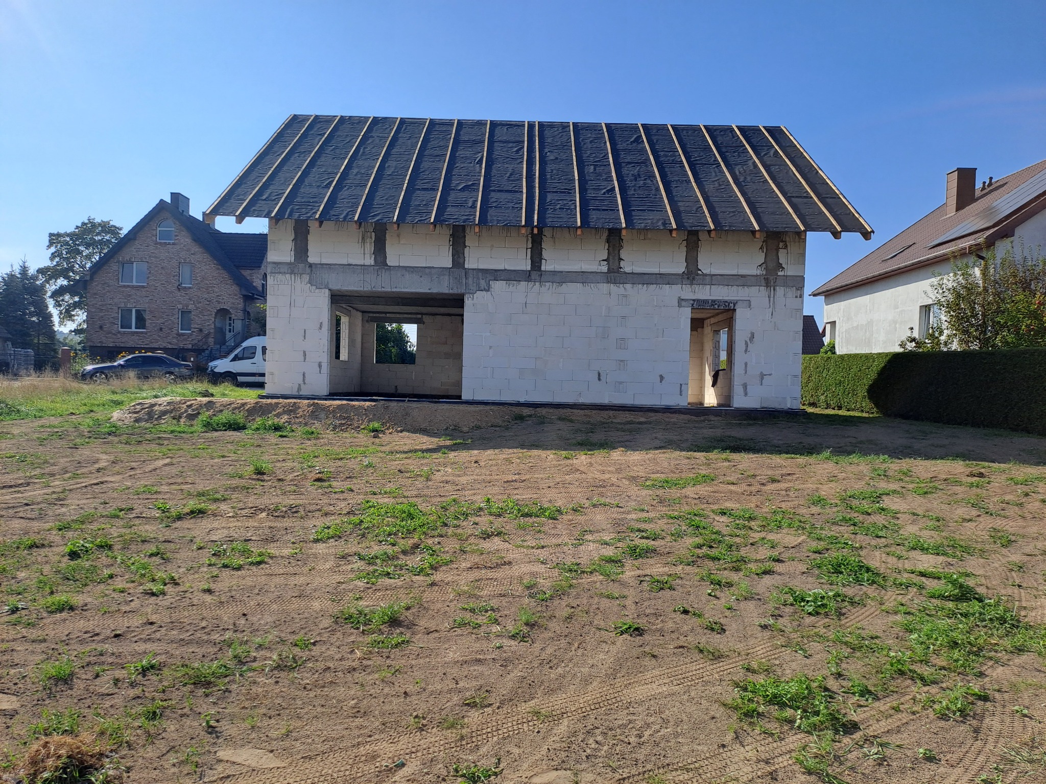 Niedokończony dom z białego pustaka, z czarną membraną na dachu, na tle zieleni i błękitnego nieba. Widoczne otwory okienne i drzwiowe, teren wokół domu niezagospodarowany.