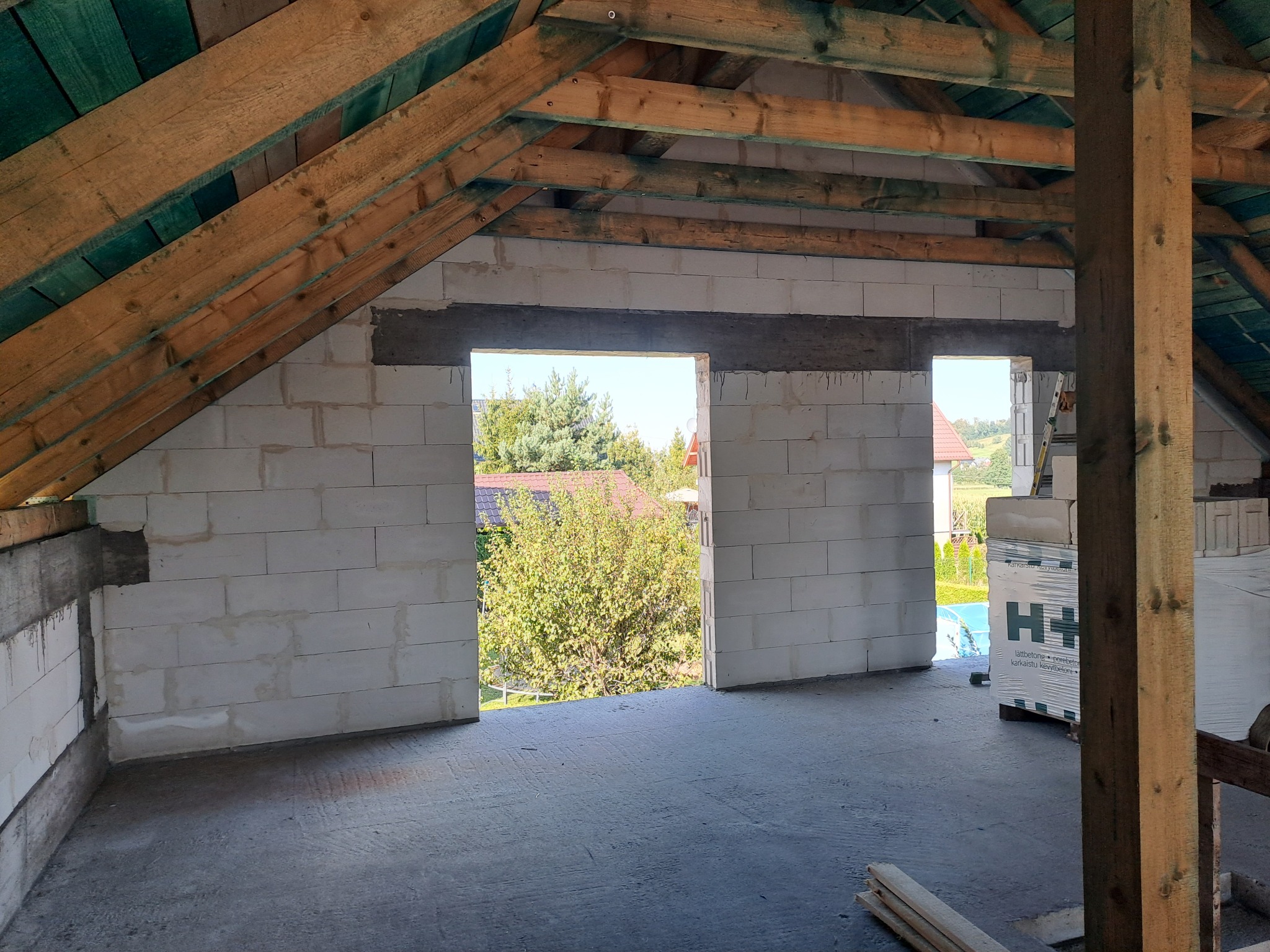 Wnętrze domu w trakcie budowy: ściany z betonu komórkowego, drewniana konstrukcja dachu, widok na zieleń przez otwory okienne. Częściowo widoczne bloczki H+H.