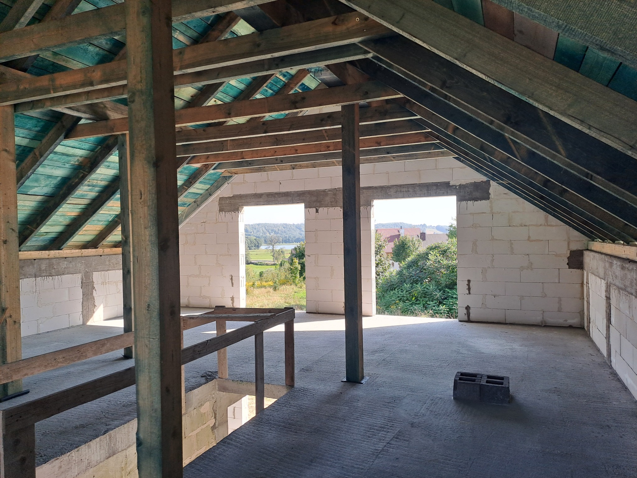 Surowy stan poddasza w domu z białego pustaka, widoczne drewniane elementy konstrukcyjne dachu i otwarte przestrzenie okienne z widokiem na zieleń.