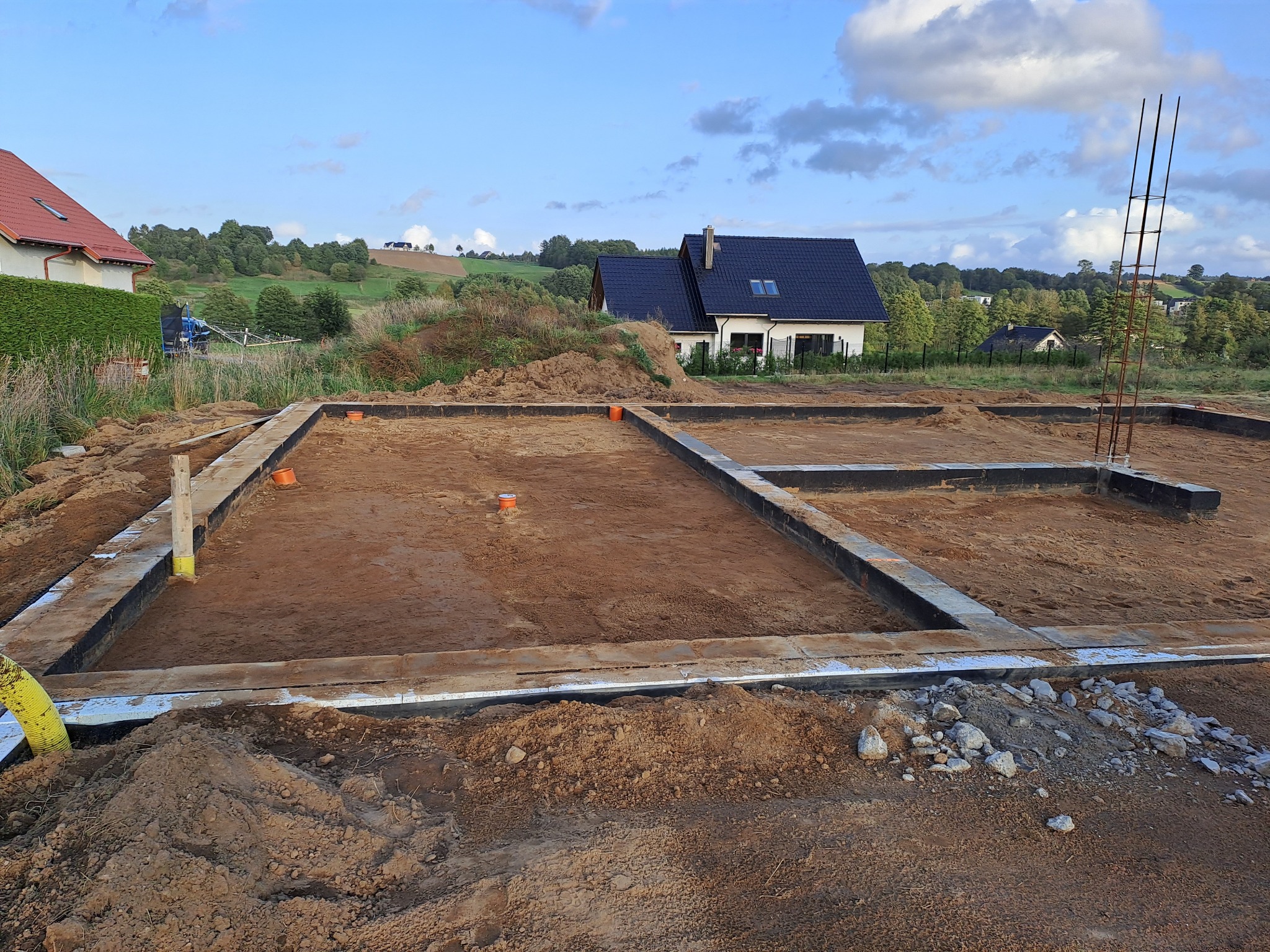 Zalane fundamenty domu jednorodzinnego z widocznym zbrojeniem i izolacją, na tle zielonego krajobrazu i błękitnego nieba. Widoczne elementy instalacji.