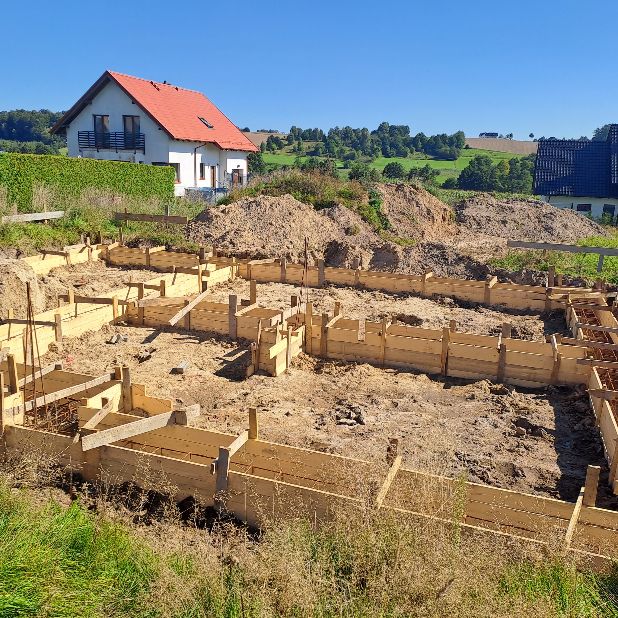 Drewniane szalunki fundamentów domu jednorodzinnego na tle pagórkowatego krajobrazu i błękitnego nieba w słoneczny dzień. Widoczne zbrojenia.