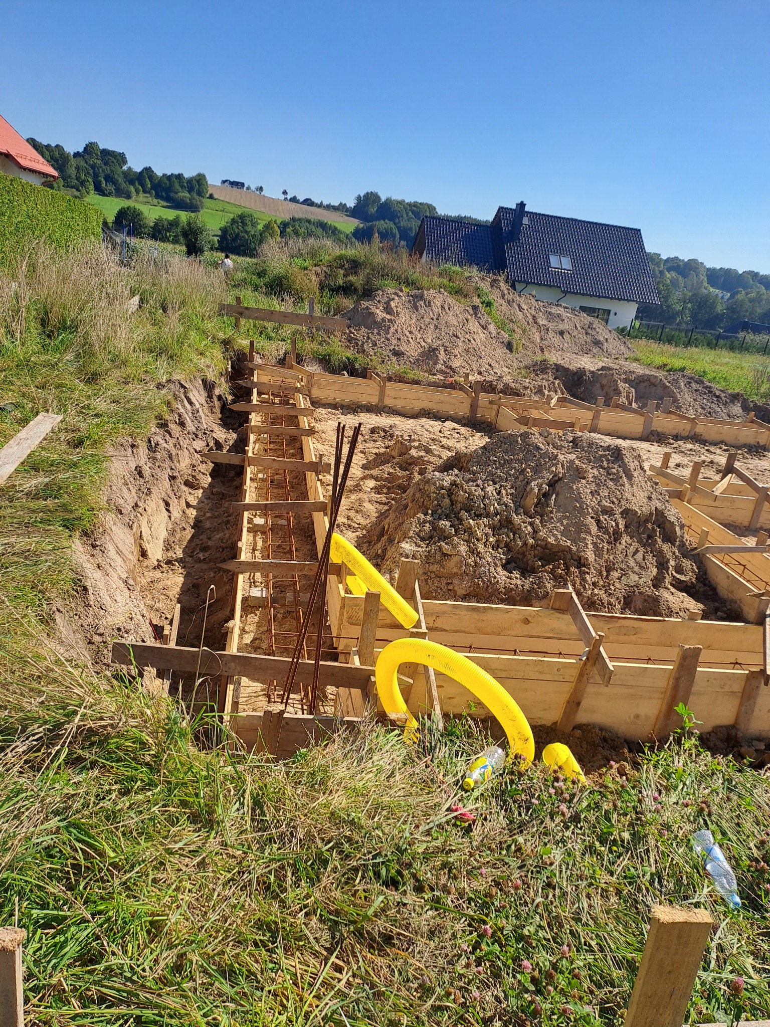 Przygotowanie szalunku i zbrojenia pod fundament domu na nierównym terenie. Widoczne żółte rury instalacyjne i drewniane elementy konstrukcyjne.