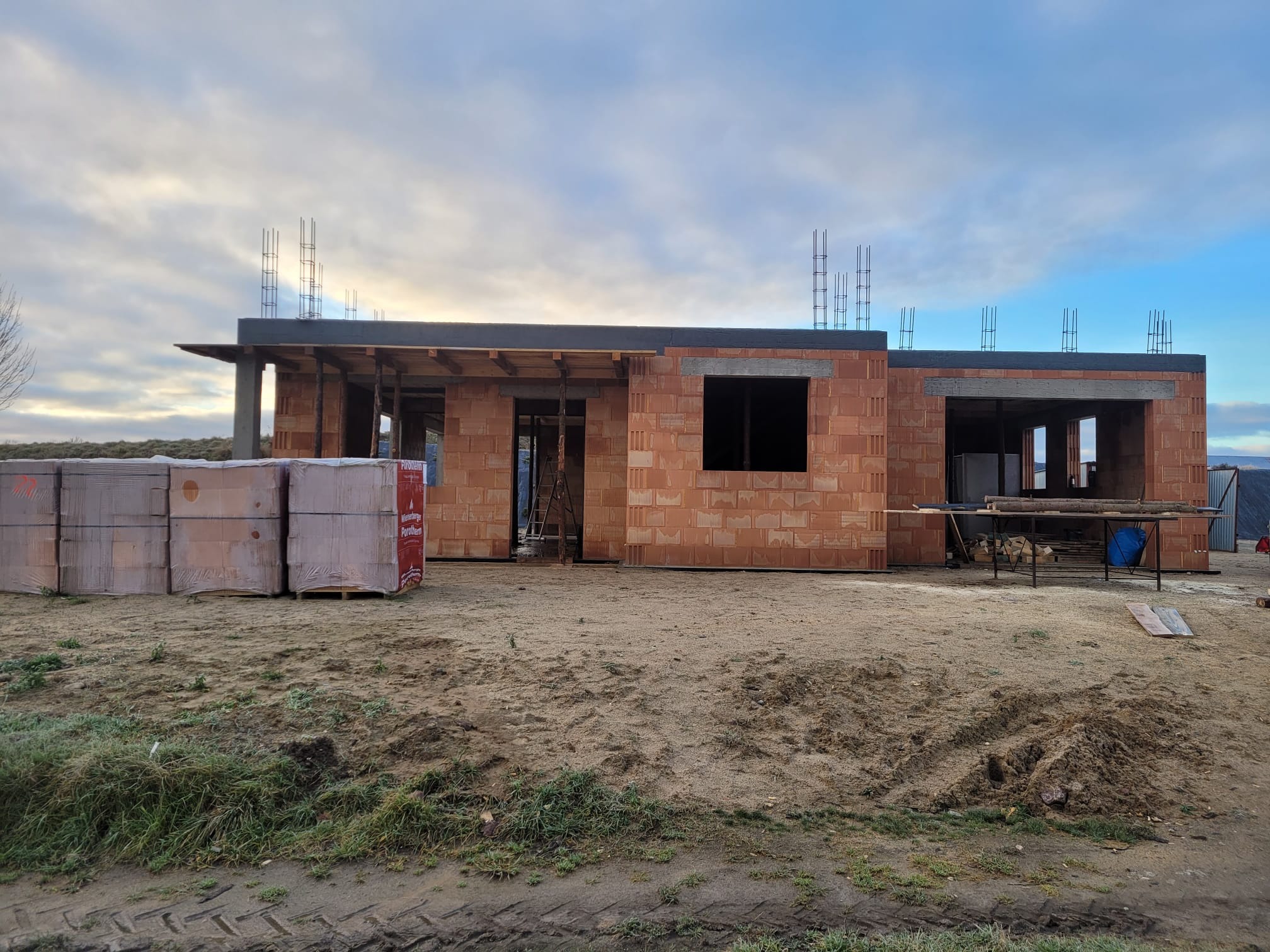 Surowy stan domu z czerwonej cegły, widoczne zbrojenia na dachu, palety z cegłami na pierwszym planie, ziemiste podłoże, pochmurne niebo w tle.