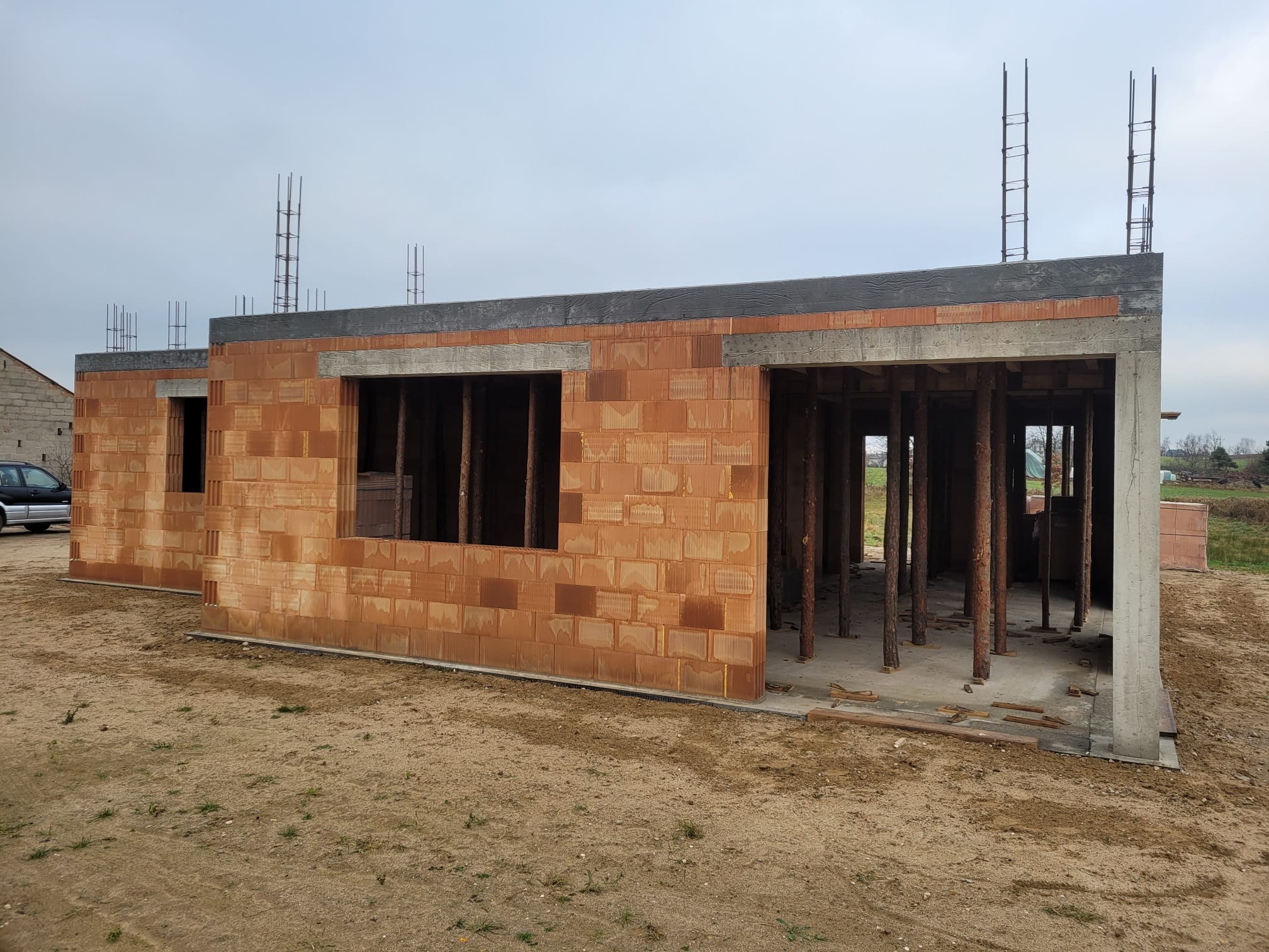 Surowy stan domu z czerwonej cegły, widoczne zbrojenia i drewniane podpory, na piaszczystym podłożu. Budowa w toku, bez wykończenia.