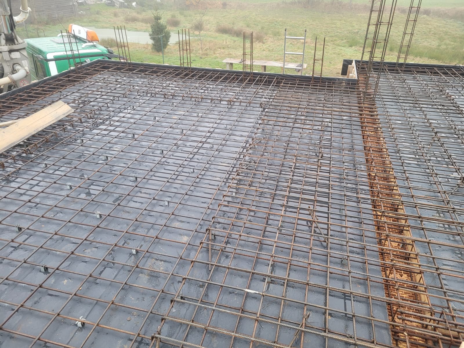 Zbrojenie stropu na czarnej folii, widok z góry. W tle zielony pojazd budowlany i drabina. Elementy konstrukcyjne przygotowane do zalania betonem.