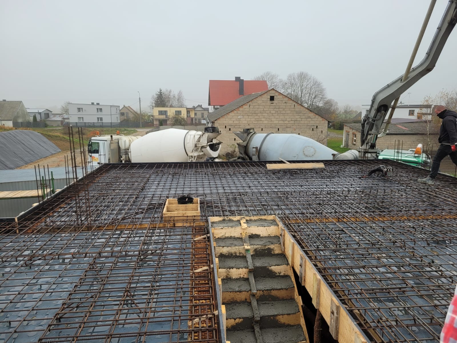 Zbrojenie stropu z betonem w szalunku, widoczne dwa betoniarki i dźwig podający beton. W tle zabudowa wiejska i pochmurne niebo. Etap budowy domu.
