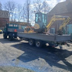 FHU Mateusz Frąckowiak - Transport koparki AMMANN YANMAR na platformie ciężarówki Unimog. Sprzęt budowlany gotowy do pracy na tle zabudowań gospodarczych w słoneczny dzień.