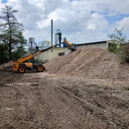 FHU Mateusz Frąckowiak - Żółta ładowarka teleskopowa JCB nabiera gruz na tle budynku z cegły i pochmurnego nieba. Widoczne ślady opon na ziemi.