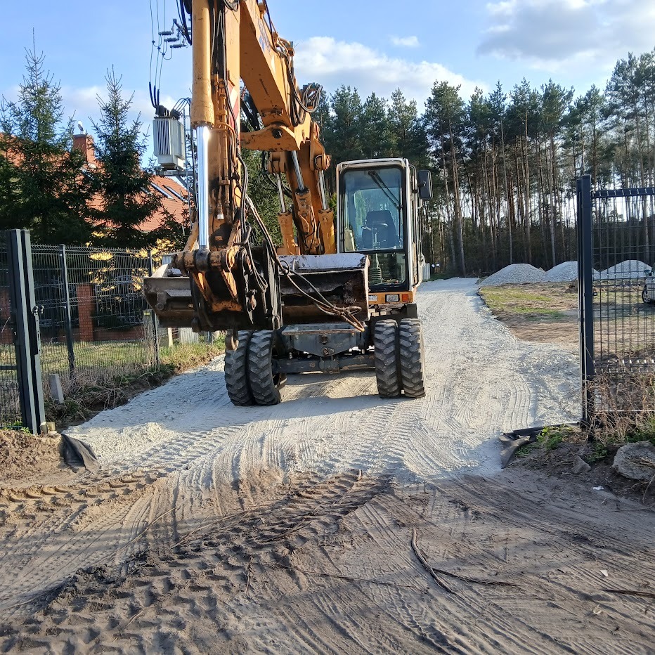 Żółta koparka na utwardzonej, szarej drodze wjazdowej, widok zza bramy. W tle las i dom. Ślady opon na piasku. Błękitne niebo z chmurami.