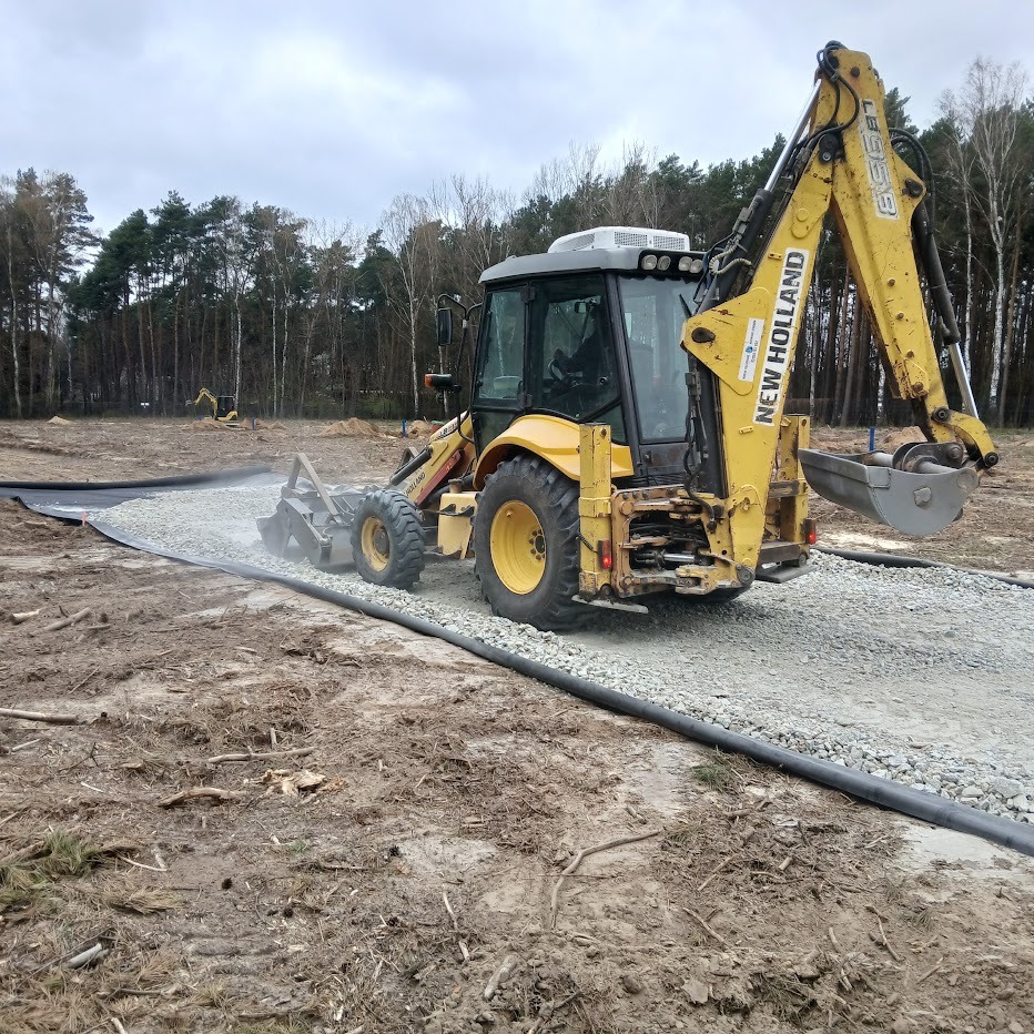 Żółta koparko-ładowarka New Holland podczas utwardzania drogi żwirowej na czarnej geowłókninie, widok z tyłu, las w tle, pochmurne niebo.