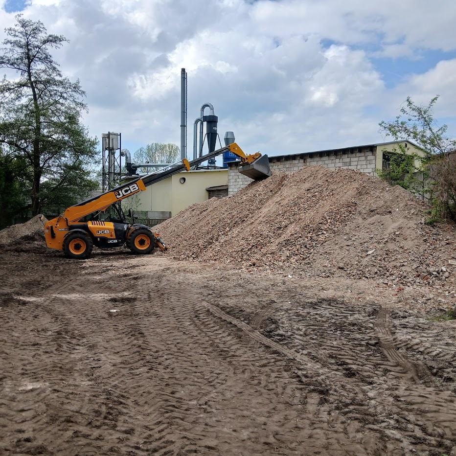Żółta ładowarka teleskopowa JCB nabiera gruz na tle budynku z cegły i pochmurnego nieba. Widoczne ślady opon na ziemi.