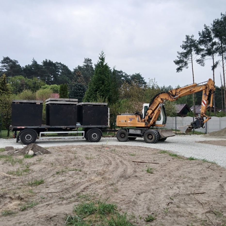 Żółta koparka kołowa Liebherr i przyczepa z trzema betonowymi szambami na żwirowej drodze, w tle zieleń i szary betonowy płot.