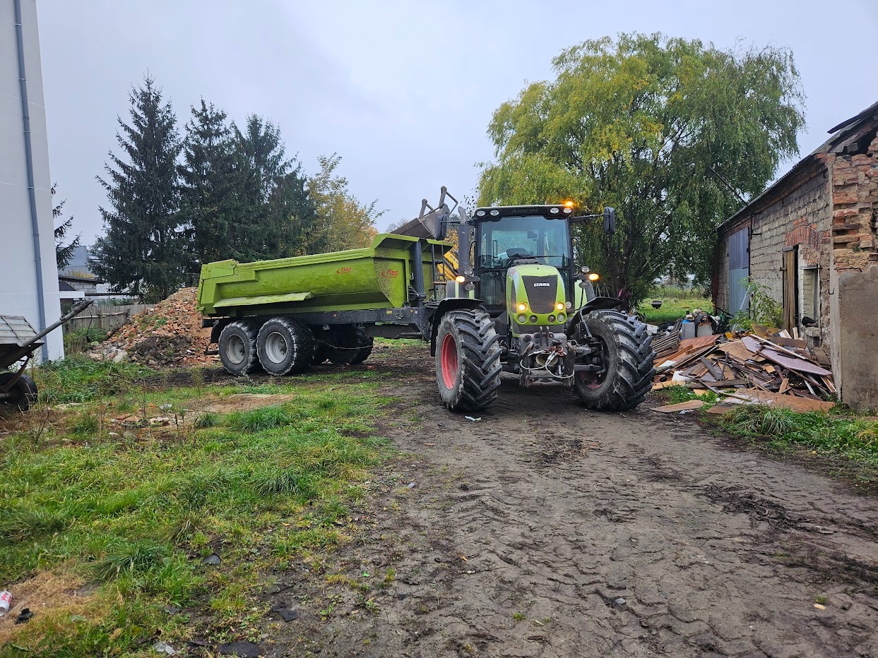 Ciągnik rolniczy z przyczepą wywrotką, załadowaną gruzem, na błotnistym terenie budowy. W tle budynek z cegły i drzewa.