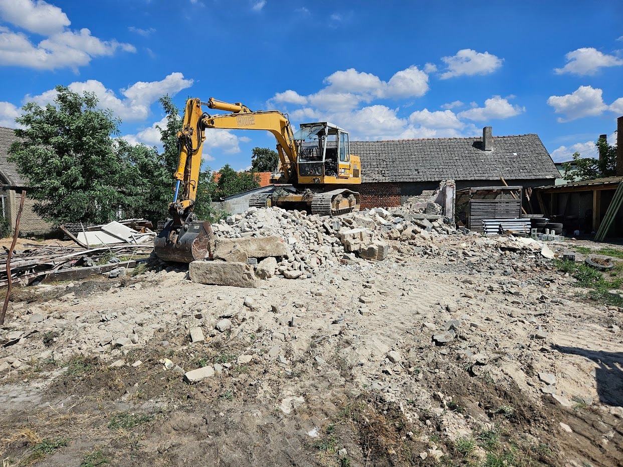 Żółta koparka na gruzowisku po rozbiórce budynku, z fragmentami murów i dachem w tle, pod błękitnym niebem z chmurami. Widoczne ślady opon na ziemi.