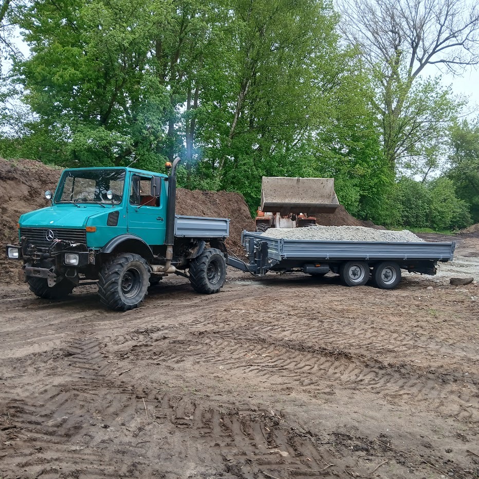 Niebieski Unimog z przyczepą pełną kruszywa na błotnistym terenie, w tle koparka i hałda ziemi. Widoczne ślady opon.