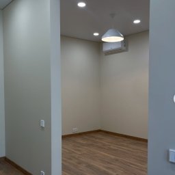 Ion popescu - Jasne wnętrze po remoncie: drewnopodobna podłoga, kremowe ściany, biała lampa wisząca i klimatyzator. Widoczne gniazdka i włączniki.